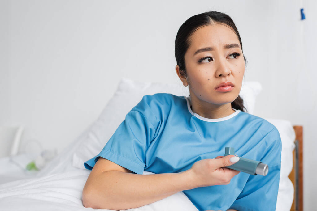 molesto y reflexivo asiático mujer sosteniendo inhalador y mirando hacia otro lado en hospital - Foto, Imagen