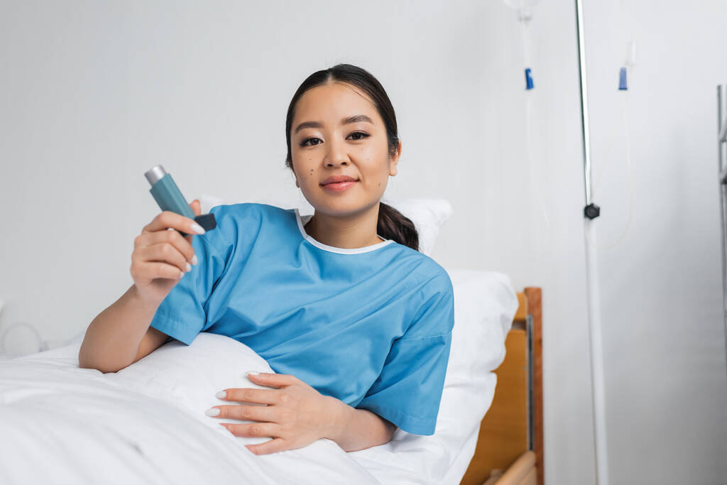 positivo asiático mujer sonriendo a cámara mientras celebración inhalador en cama en hospital sala - Foto, Imagen