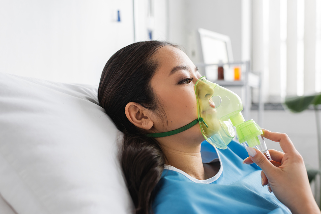 boční pohled na nemocné asijské ženy v kyslíkové masce ležící na posteli v nemocnici oddělení - Fotografie, Obrázek