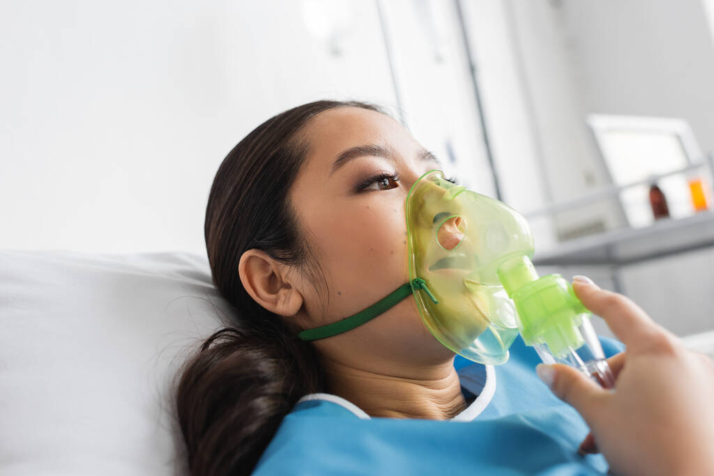 joven asiático mujer en oxígeno máscara acostado en hospital cama y mirando lejos - Foto, imagen