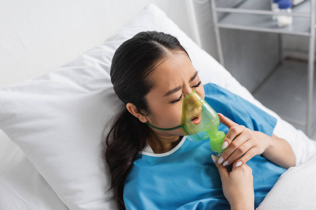 vysoký úhel pohledu na ustaranou asijskou ženu se zavřenýma očima dýchání v kyslíkové masce na nemocniční posteli - Fotografie, Obrázek