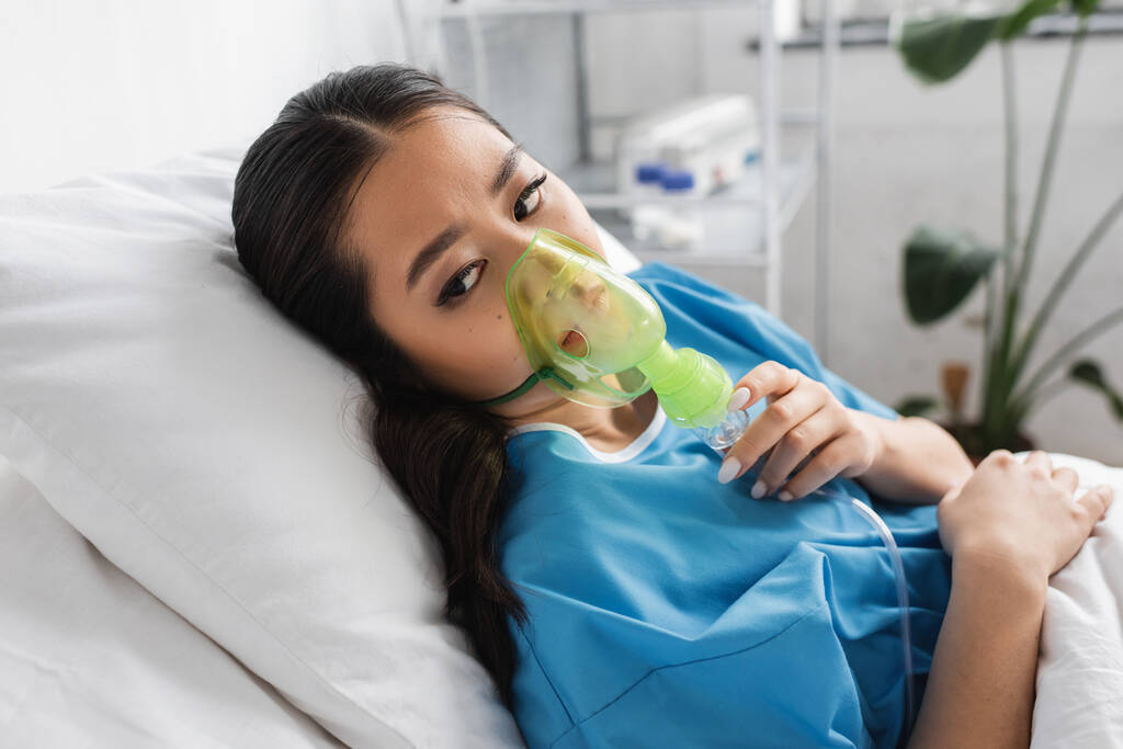 νεαρή Ασιάτισσα κοιτάζει αλλού ενώ βρίσκεται στο κρεβάτι του νοσοκομείου με μάσκα οξυγόνου - Φωτογραφία, εικόνα