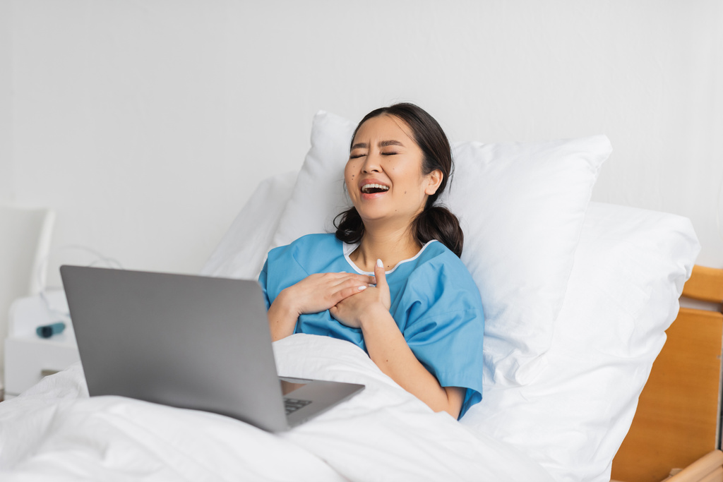 animado ásia mulher tocando peito e rindo com fechado olhos enquanto assistindo comédia filme no laptop na clínica - Foto, Imagem