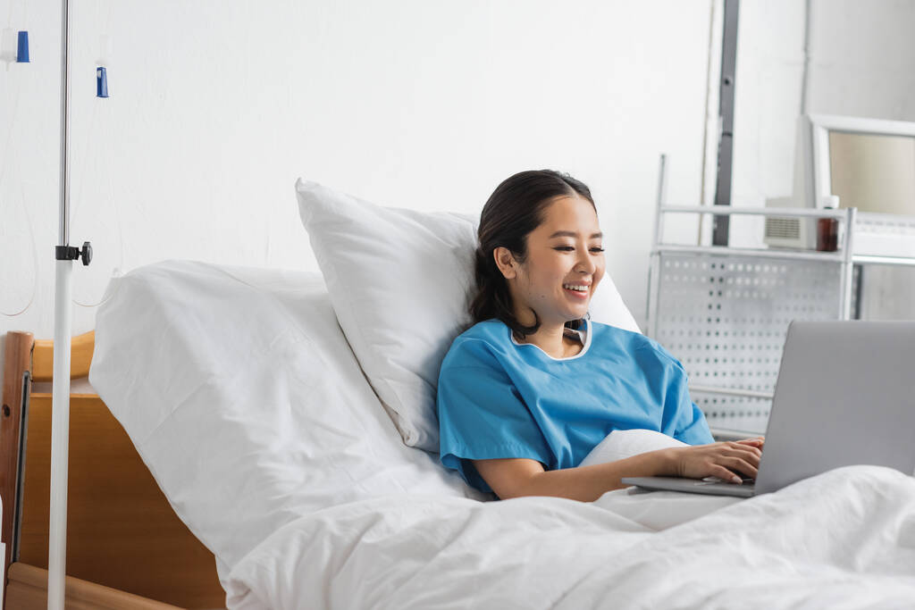 alegre asiático mujer mecanografía en portátil mientras permaneciendo en hospital cama - Foto, imagen