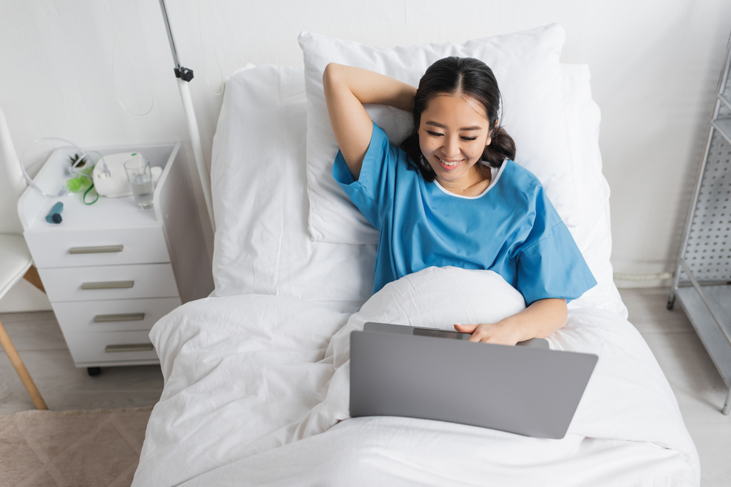 alto ângulo vista de alegre asiático mulher usando laptop no cama no clínica - Foto, Imagem