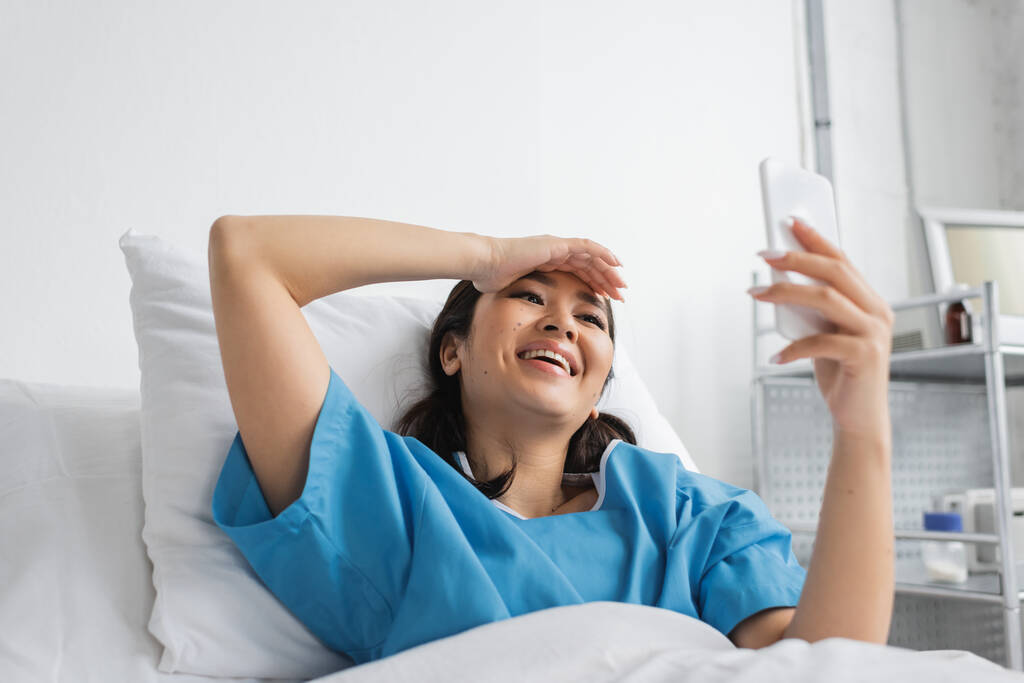 ενθουσιασμένοι ασιατική γυναίκα αγγίζοντας το μέτωπο και κοιτάζοντας smartphone στο κρεβάτι του νοσοκομείου - Φωτογραφία, εικόνα