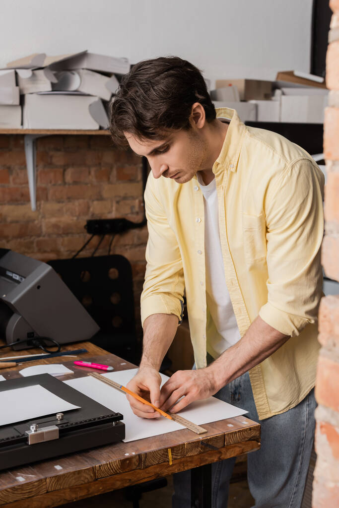 όμορφος χειριστής εκτύπωσης κρατώντας χάρακα και μολύβι κοντά στο χαρτί στο σύγχρονο κέντρο εκτύπωσης  - Φωτογραφία, εικόνα