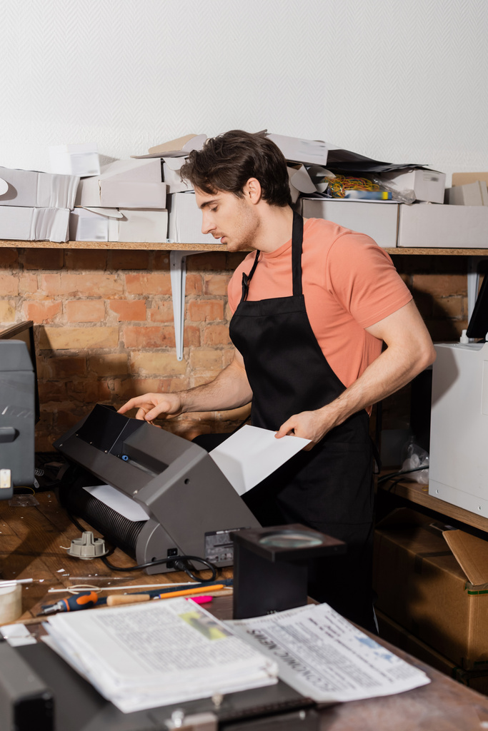 homem bonito em avental segurando papel perto de impressora e jornais no centro de impressão  - Foto, Imagem