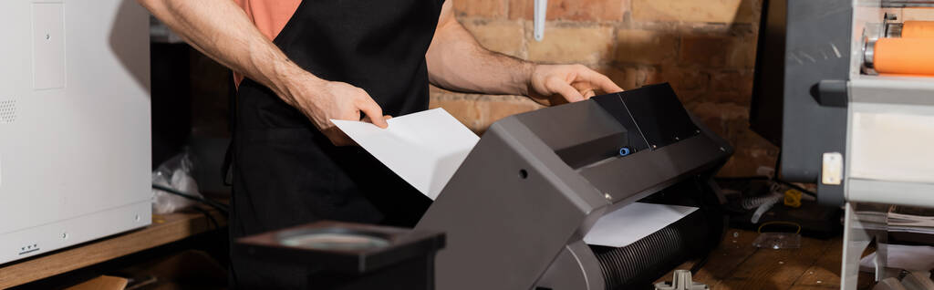 bijgesneden weergave van de werknemer in een schort papier in de buurt van de printer en verse kranten in het prentencentrum, banner  - Foto, afbeelding