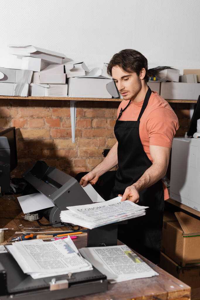 tipógrafo guapo en delantal con periódicos impresos en la sala de fotocopias  - Foto, Imagen