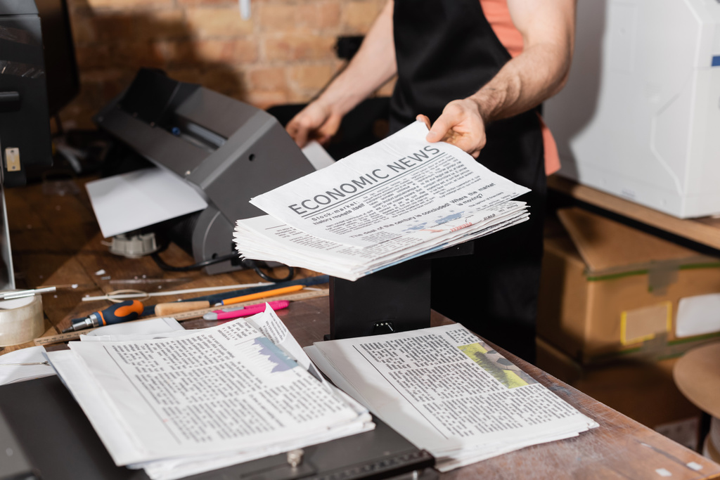 abgeschnittene Ansicht eines Typographen in Schürze, der gedruckte Zeitungen mit Wirtschaftsnachrichten hält  - Foto, Bild