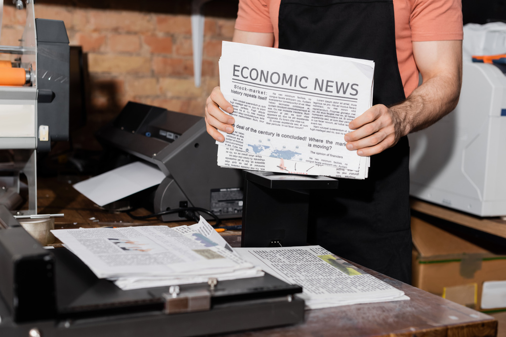bijgesneden beeld van jonge typograaf in schort met kranten met economisch nieuws   - Foto, afbeelding