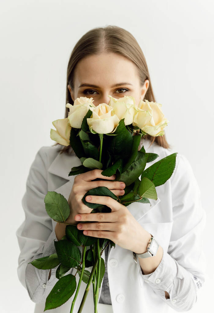молодий лікар у білому пальто тримає в руках букет білих троянд на білому тлі. дерматолог. професійна медицина
 - Фото, зображення
