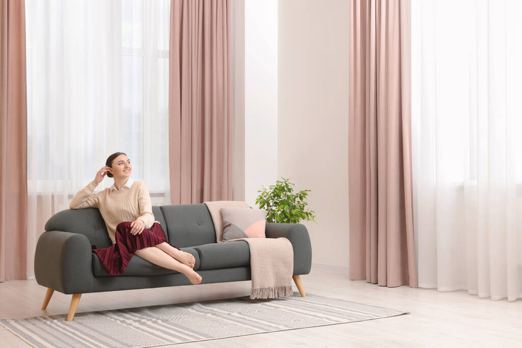 Женщина сидит на диване возле окна со стильными занавесками дома. Пространство для текста - Фото, изображение