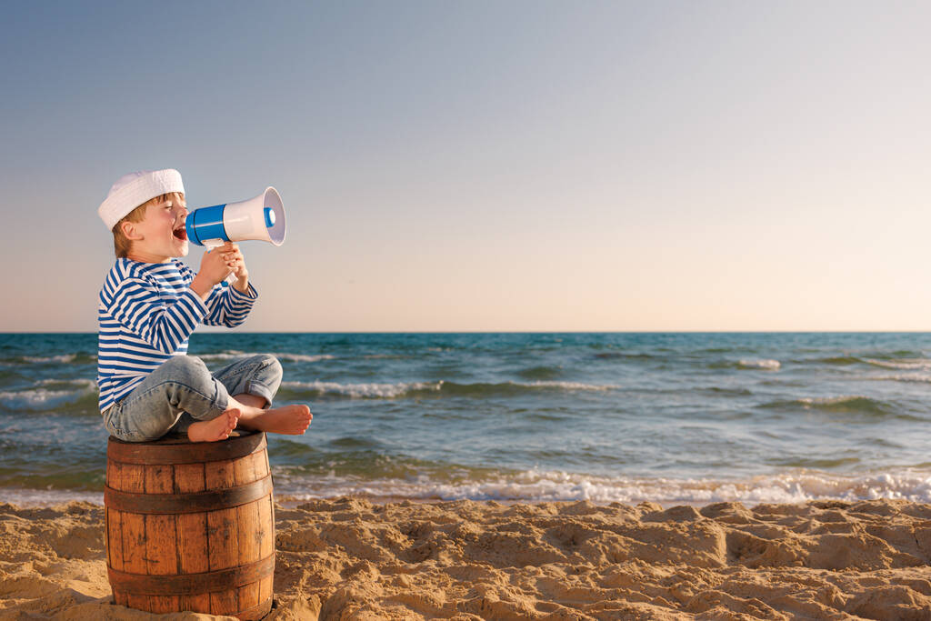 El niño finge ser marinero. El chico sostiene el altavoz. Niño sentado en un viejo barril en la playa. Un chico de vacaciones de verano. Concepto de aventura y viajes - Foto, imagen