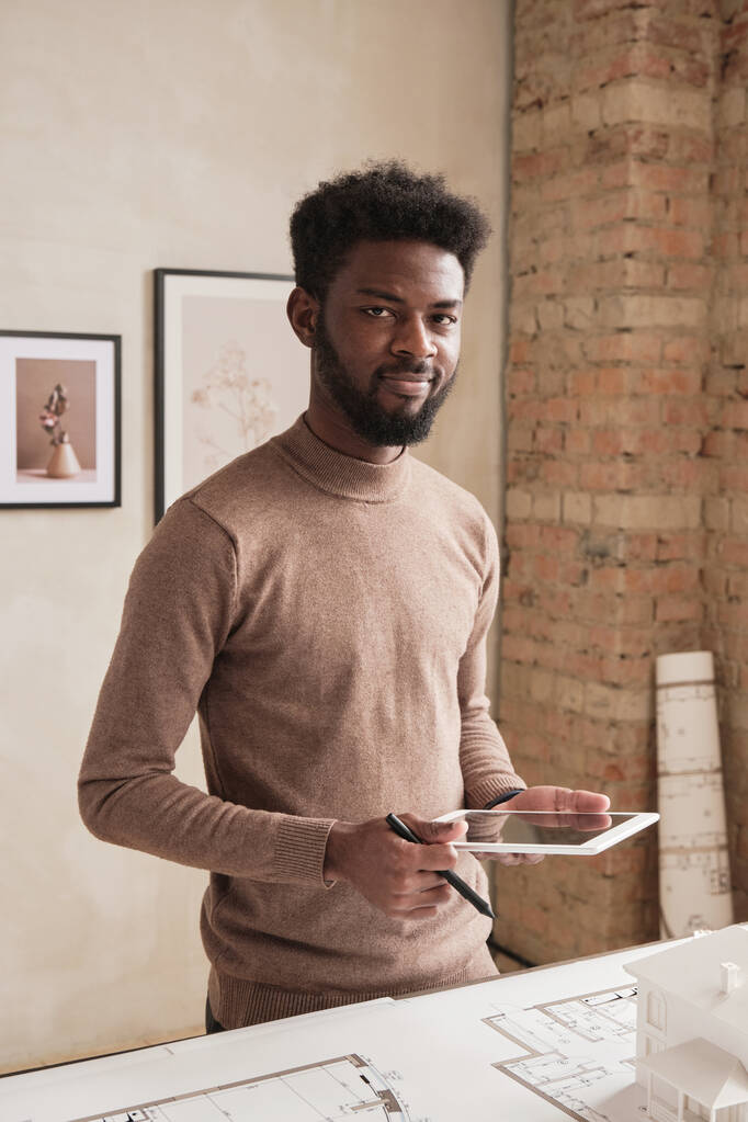 Retrato de joven arquitecto negro sonriente con barba de pie en la mesa con plano y usando tableta mientras se elabora el diseño del edificio - Foto, imagen