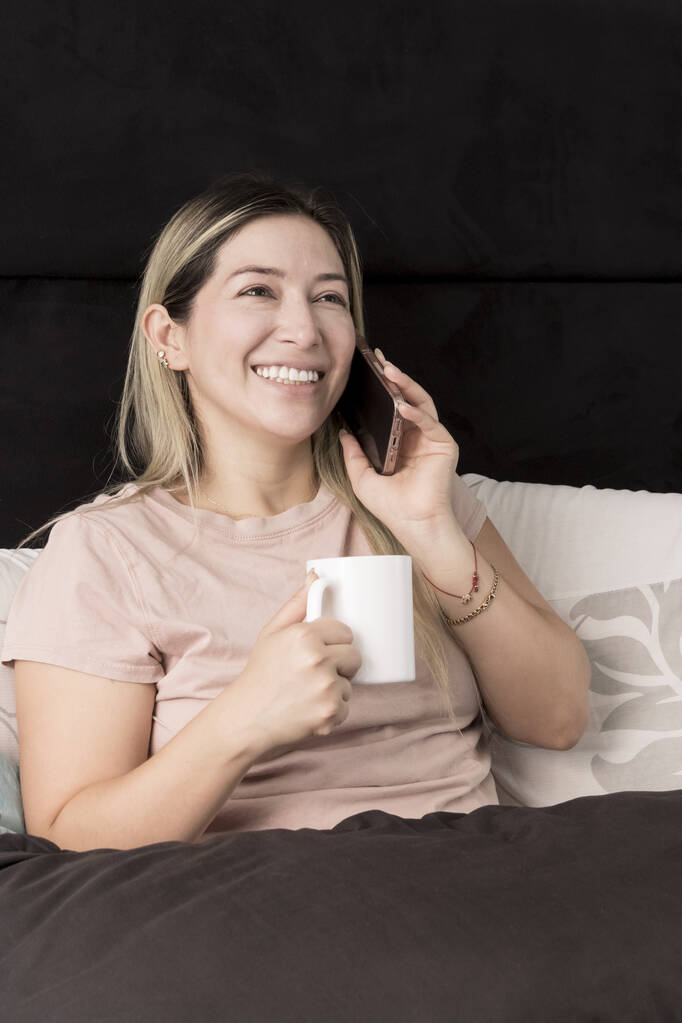 Plusz méretű lány beszél mobilon az ágyában, miközben kávét iszik. - Fotó, kép