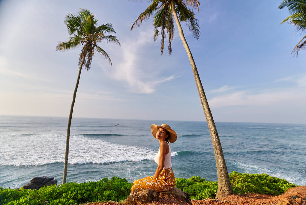 Güneş doğarken avuçlar arasında okyanus manzaralı tropik bir yerde renkli giysiler içinde poz veren Afrikalı genç bir kadın model. Şafak vakti deniz manzaralı, palmiye ağaçları olan egzotik manzaraya karşı oturan siyah kadın. - Fotoğraf, Görsel