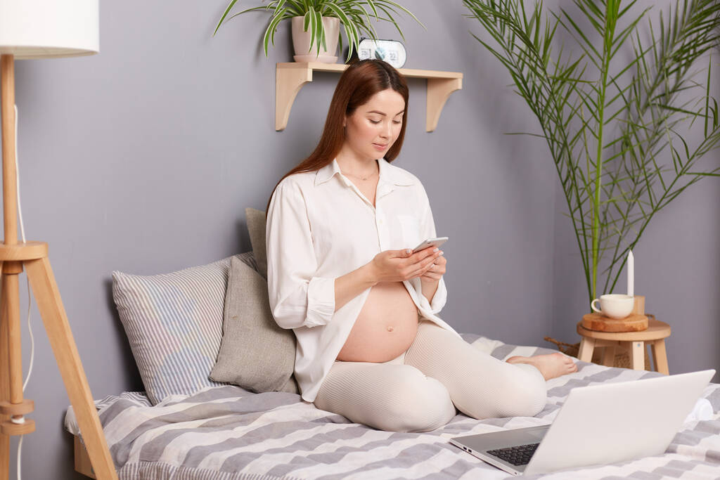 Ziemlich positive schwangere braunhaarige Kaukasierin sitzt im Bett und benutzt ihr Handy zum Tippen von Nachrichten und ihren Laptop für die Online-Arbeit. - Foto, Bild