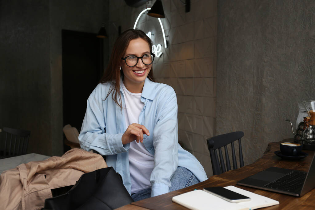 Πορτρέτο της νεαρής όμορφης γυναίκας ντυμένη με έξυπνο casual στυλ κάθεται σε μια καφετέρια με το laptop της ένα φλιτζάνι ζεστό ρόφημα. Σχεδίαση απομακρυσμένης εργασίας. Κλείσιμο, αντιγραφή χώρου, φόντο. - Φωτογραφία, εικόνα
