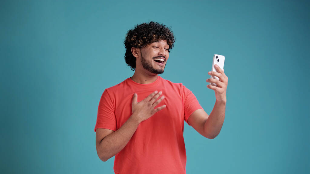 Νέος όμορφος χαμογελαστός Ινδός κάνει βιντεοκλήση στο smartphone, χαιρετώντας, συνομιλώντας μέσω κινητού τηλεφώνου απομονωμένο σε μπλε φόντο στούντιο. - Φωτογραφία, εικόνα