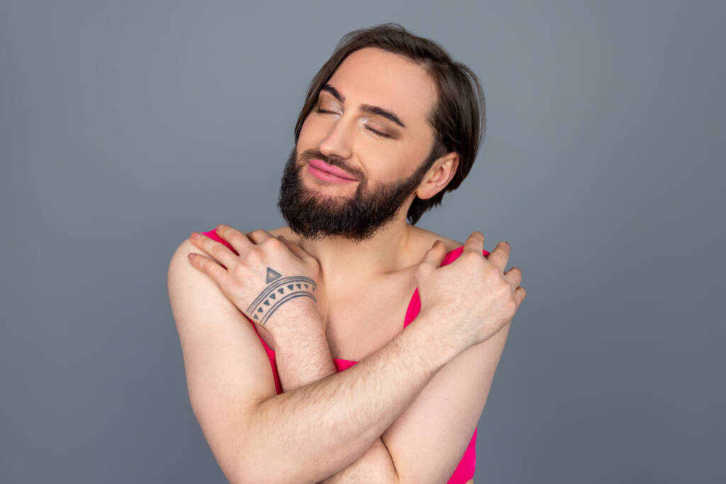 Zufriedene selbstverliebte Brünette Transsexuelle tragen rosa Top umarmt sich mit Lächeln und Freude posiert isoliert vor grauem Hintergrund - Foto, Bild