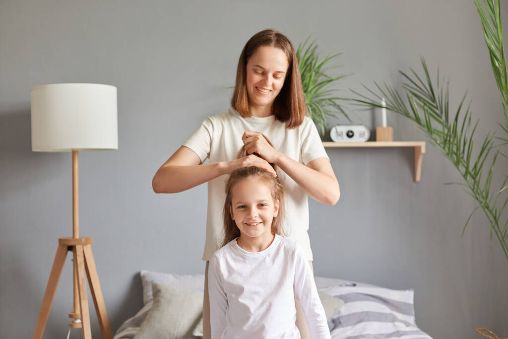 Positive cheerful mother making hairstyle to her daughter while they sitting on bed in bedroom, doing ponytail to kid before going to school in the morning. - Photo, Image