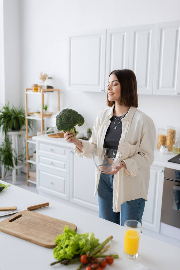 Lächelnde Frau hält Brokkoli und Schüssel in der Nähe von Gemüse und Schneidebrett in der Küche  - Foto, Bild