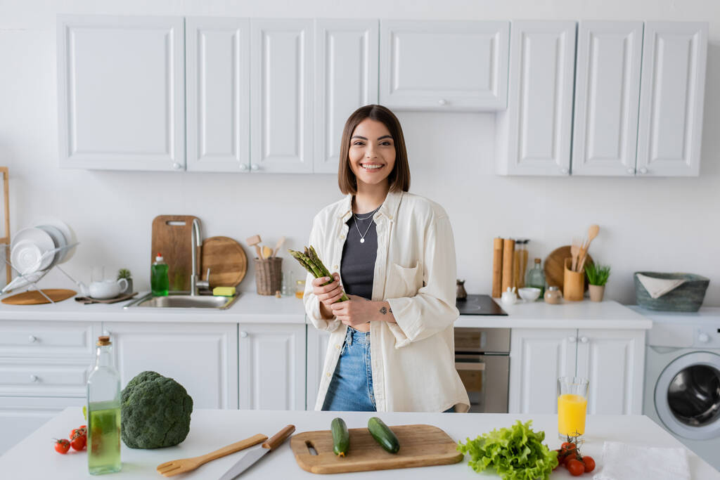 Cheerful woman holding asparagus and looking at camera near vegetables in kitchen  - Photo, Image