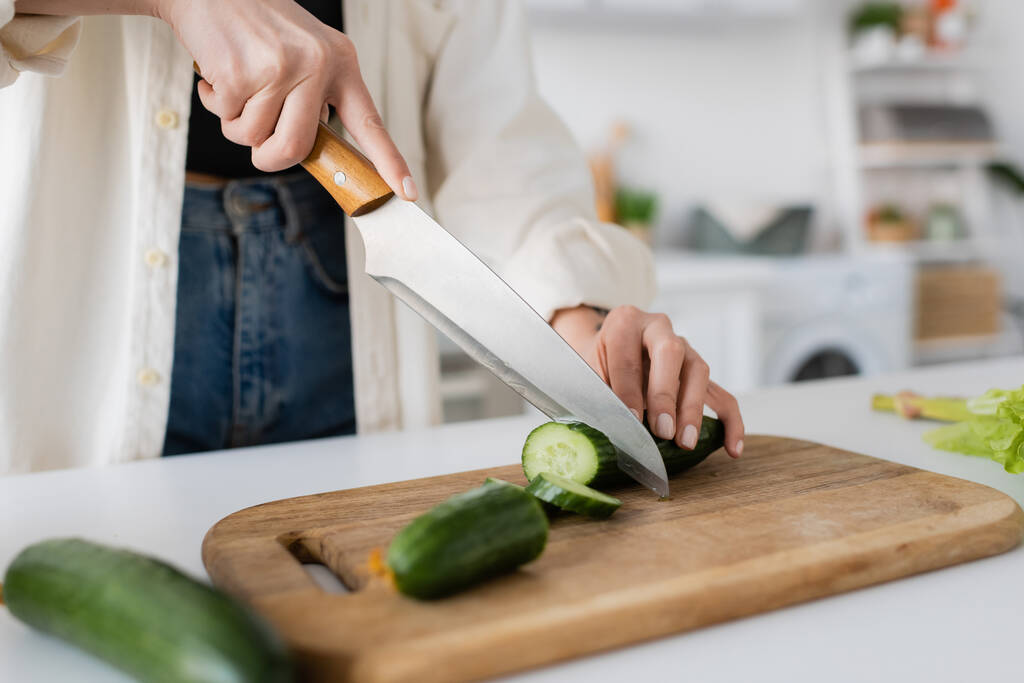 Cropped view of woman cutting ripe cucumber on chopping board in kitchen  - Photo, Image