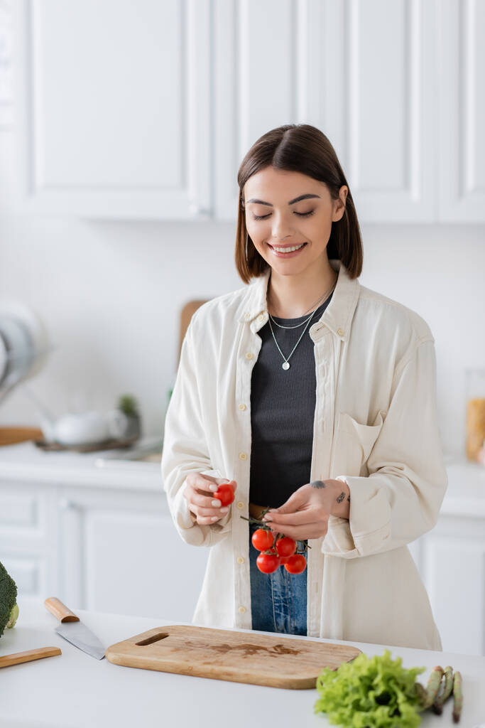 Mujer positiva sosteniendo tomates cherry cerca de verduras y tabla de cortar en la cocina  - Foto, imagen