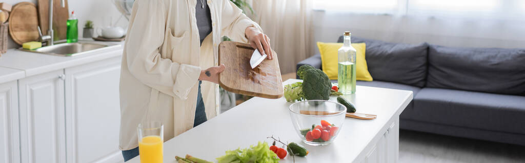 Gewassen uitzicht op getatoeëerde vrouw met mes en snijplank tijdens het koken van verse salade in de keuken, banner  - Foto, afbeelding
