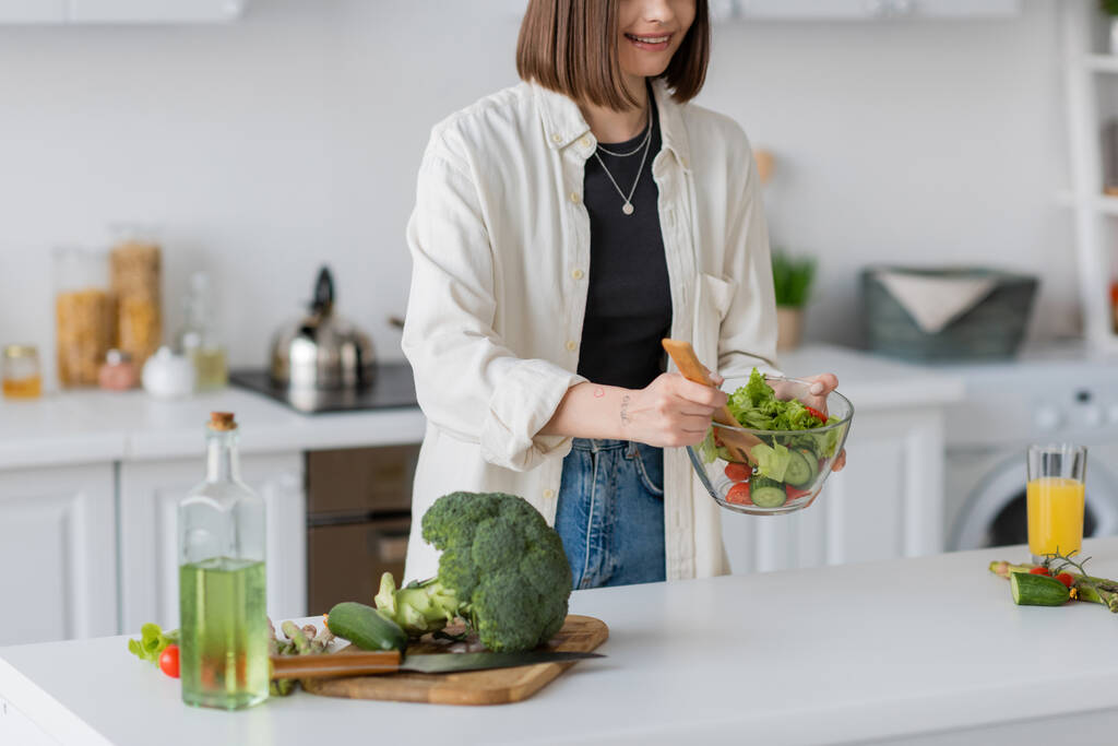 Ausgeschnittene Ansicht einer fröhlichen Frau mit Schüssel und frischem Salat in der Küche  - Foto, Bild