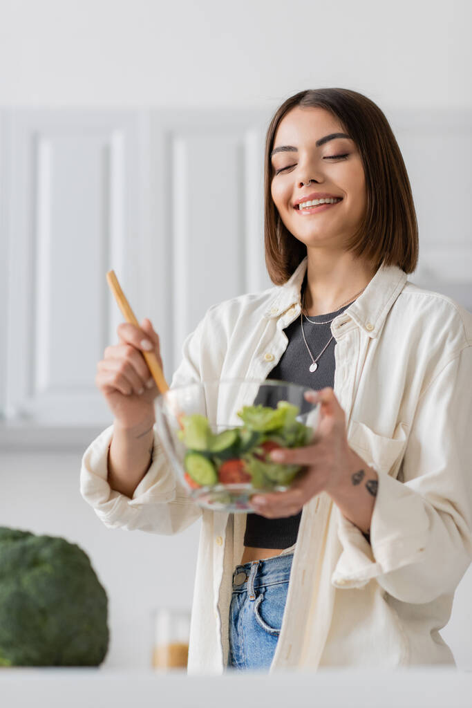 Fröhlich tätowierte Frau mixt in der heimischen Küche frischen Salat  - Foto, Bild