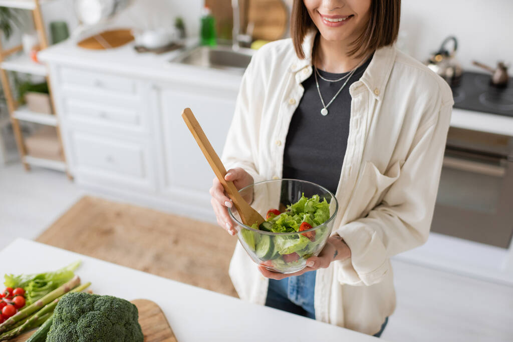 Καλλιεργημένη θέα χαμογελαστή γυναίκα κρατώντας φρέσκια σαλάτα σε μπολ στην κουζίνα  - Φωτογραφία, εικόνα