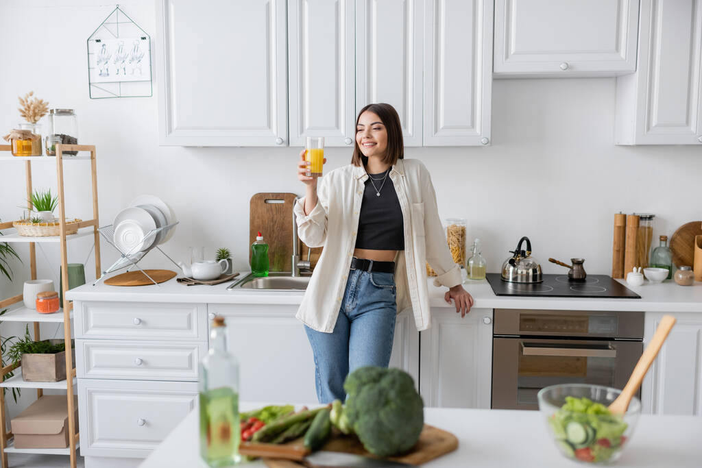 Uśmiechnięta brunetka trzyma sok pomarańczowy blisko rozmytej sałatki w kuchni  - Zdjęcie, obraz