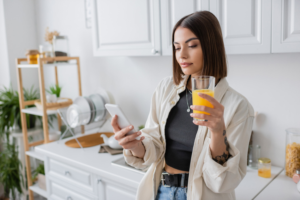 オレンジジュースを保持し、キッチンでスマートフォンを使用しているブルネットの女性  - 写真・画像