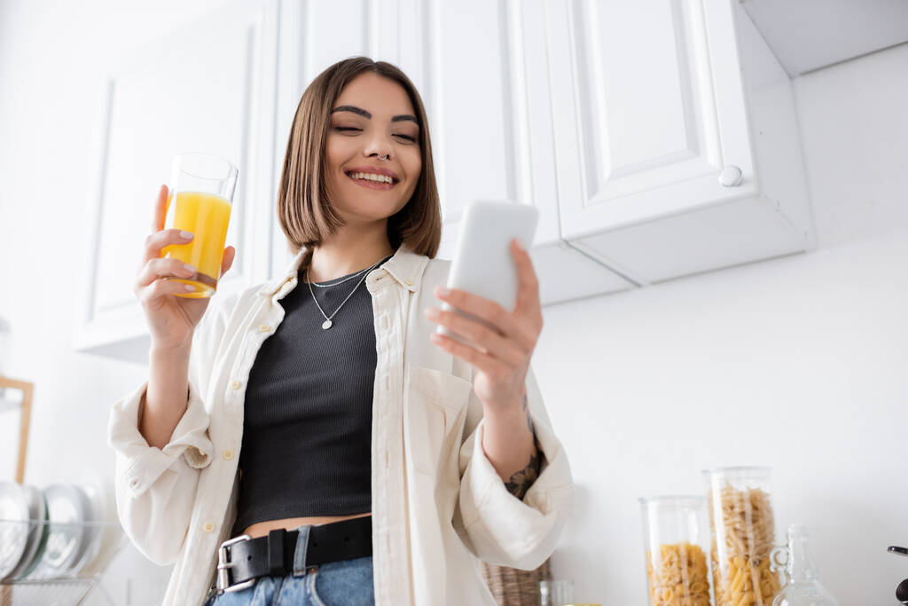 Низький кут зору усміхненої жінки, використовуючи смартфон і тримаючи апельсиновий сік на кухні
  - Фото, зображення