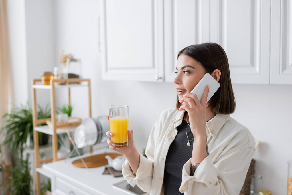 Jeune femme parlant sur smartphone et tenant du jus d'orange dans la cuisine  - Photo, image