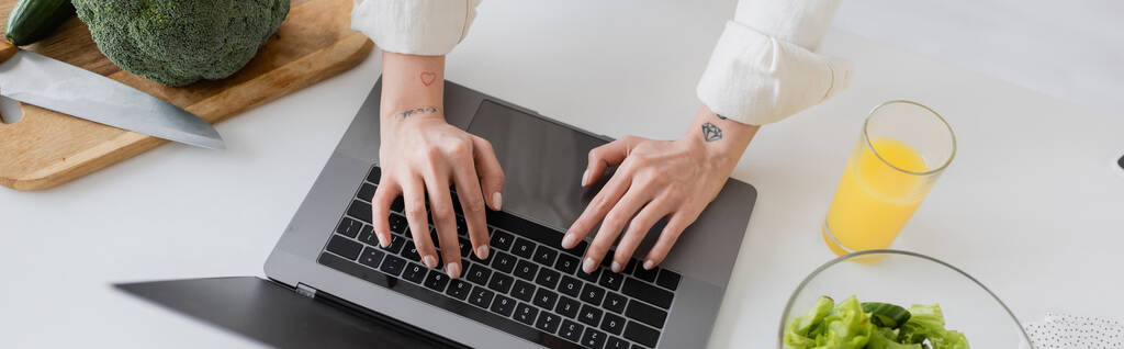 Vista recortada del freelancer tatuado usando el ordenador portátil cerca del jugo de naranja y la ensalada en la cocina, pancarta  - Foto, Imagen