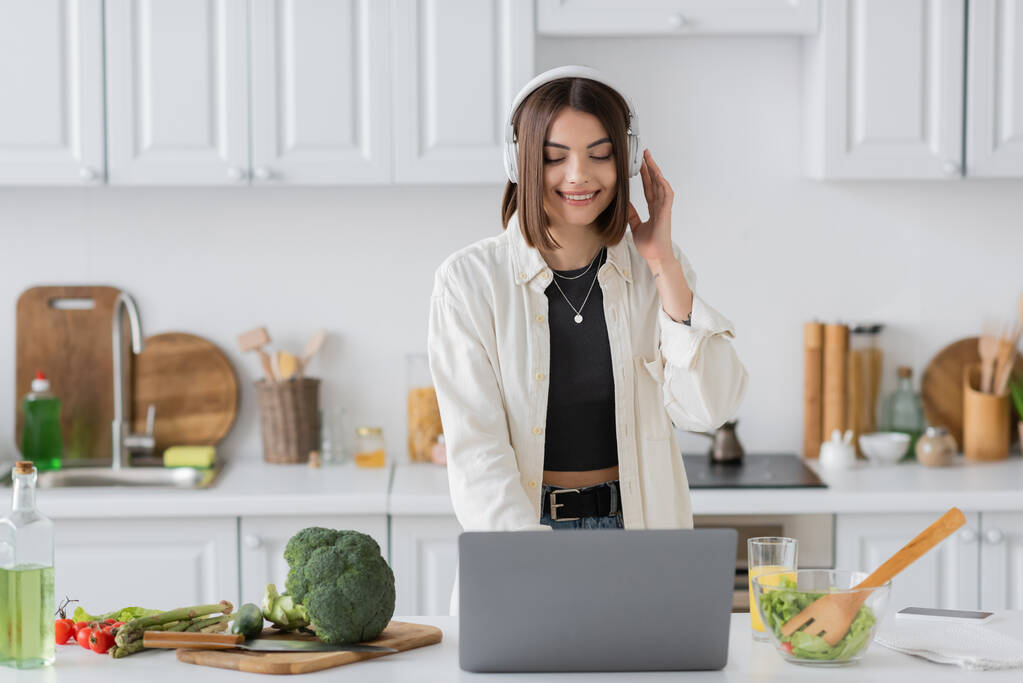 Веселая молодая женщина в наушниках с ноутбуком рядом с салатом и свежими овощами на кухне  - Фото, изображение