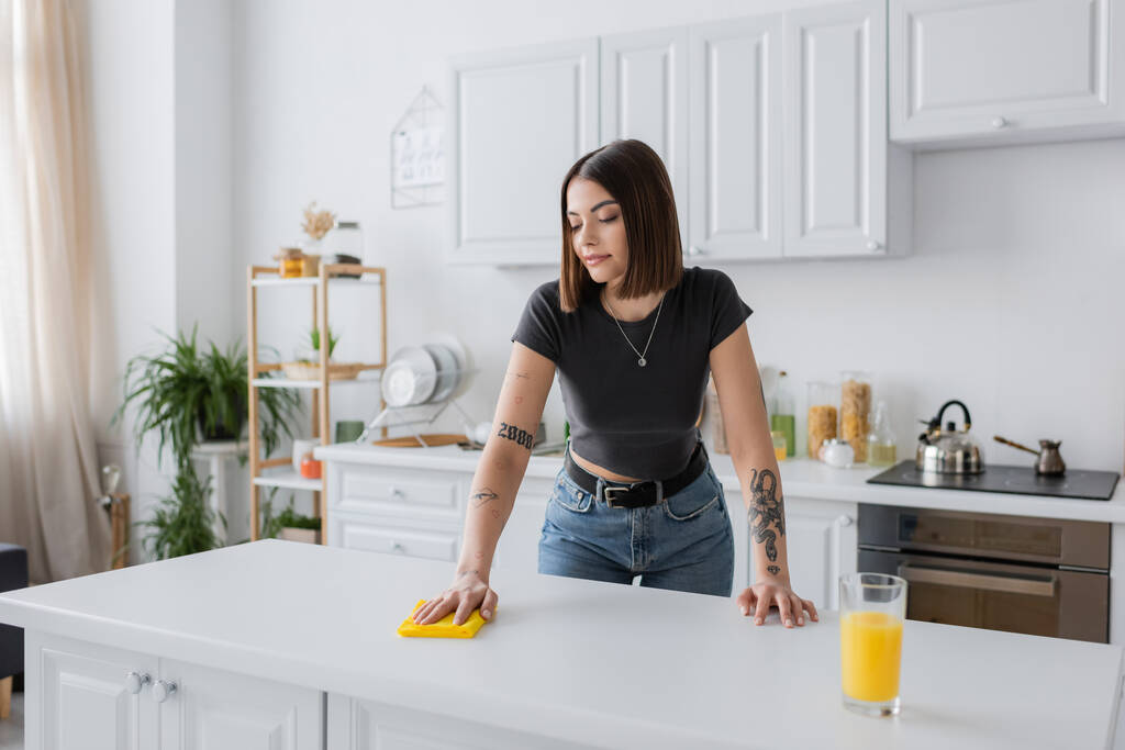 Tätowierte brünette Frau putzt Arbeitsplatte mit Lappen in Küche  - Foto, Bild
