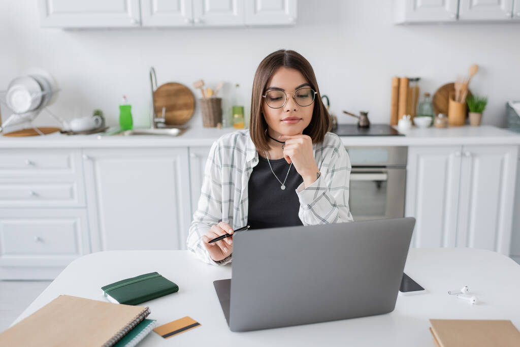 Morena freelancer mirando portátil cerca de cuadernos y tarjeta de crédito en la cocina  - Foto, imagen