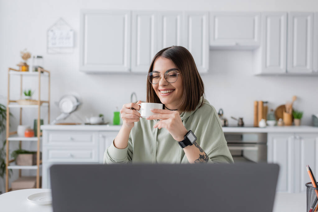 Freelancer positivo em óculos segurando café perto de laptop borrado em casa  - Foto, Imagem