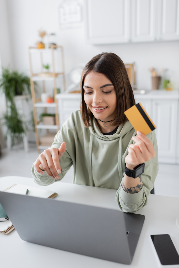 Femme positive tenant la carte de crédit pendant les achats en ligne sur ordinateur portable à la maison  - Photo, image