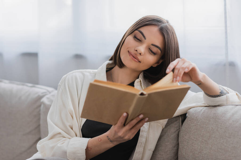Potěšená tetovaná žena čte rozmazanou knihu v obývacím pokoji  - Fotografie, Obrázek