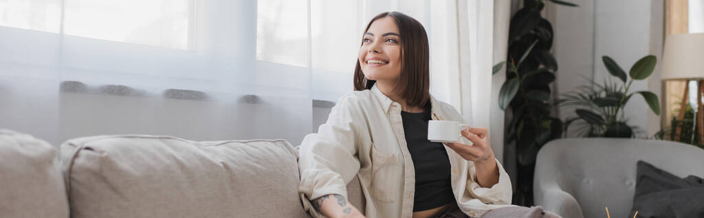 Overjoyed woman holding cup of coffee while sitting on couch at home, banner  - Photo, Image