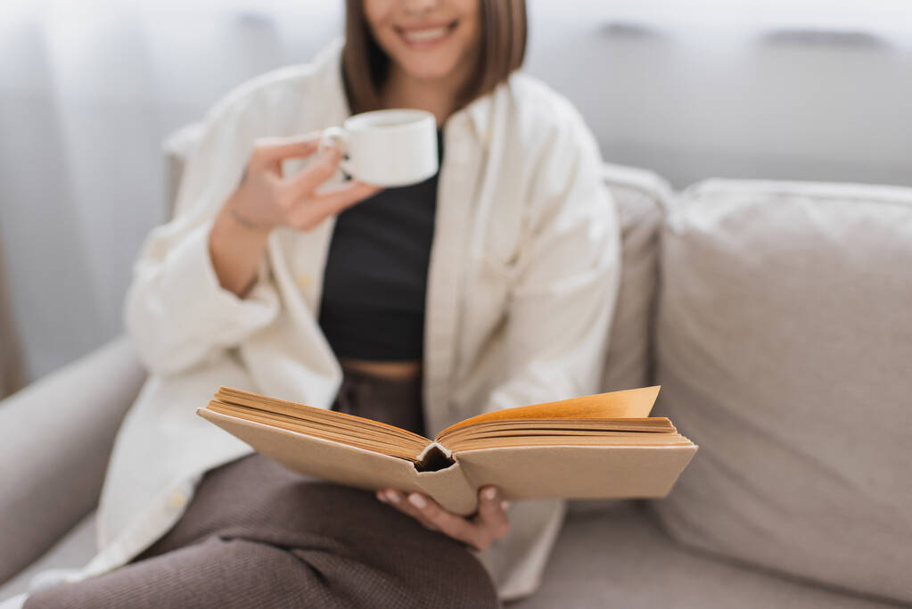 Ausgeschnittene Ansicht einer verschwommenen Frau mit Kaffeetasse und Lesebuch auf der heimischen Couch  - Foto, Bild