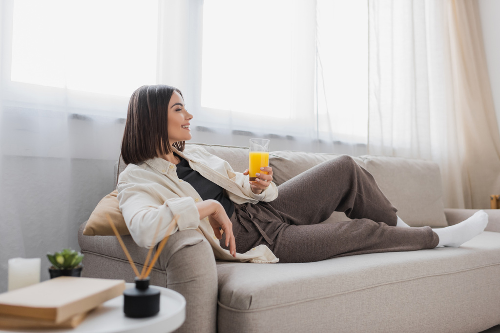 Seitenansicht einer lächelnden brünetten Frau mit einem Glas Orangensaft, während sie zu Hause auf der Couch sitzt  - Foto, Bild