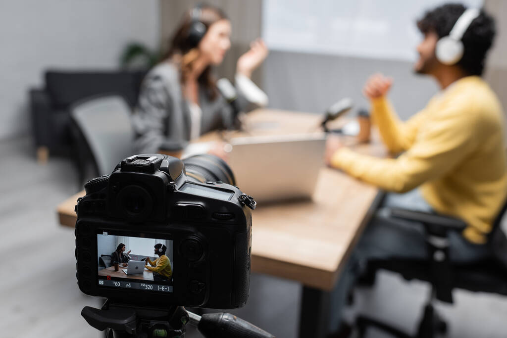 selectieve focus van professionele digitale camera opname hoe interviewer praten met jonge Indiase gast terwijl samen zitten in de buurt van laptop en microfoons op wazige achtergrond in de studio - Foto, afbeelding